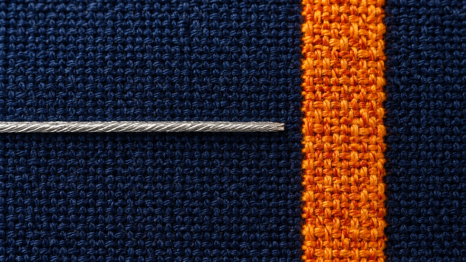 A silver thread crossing indigo fabric beside an orange woven band
