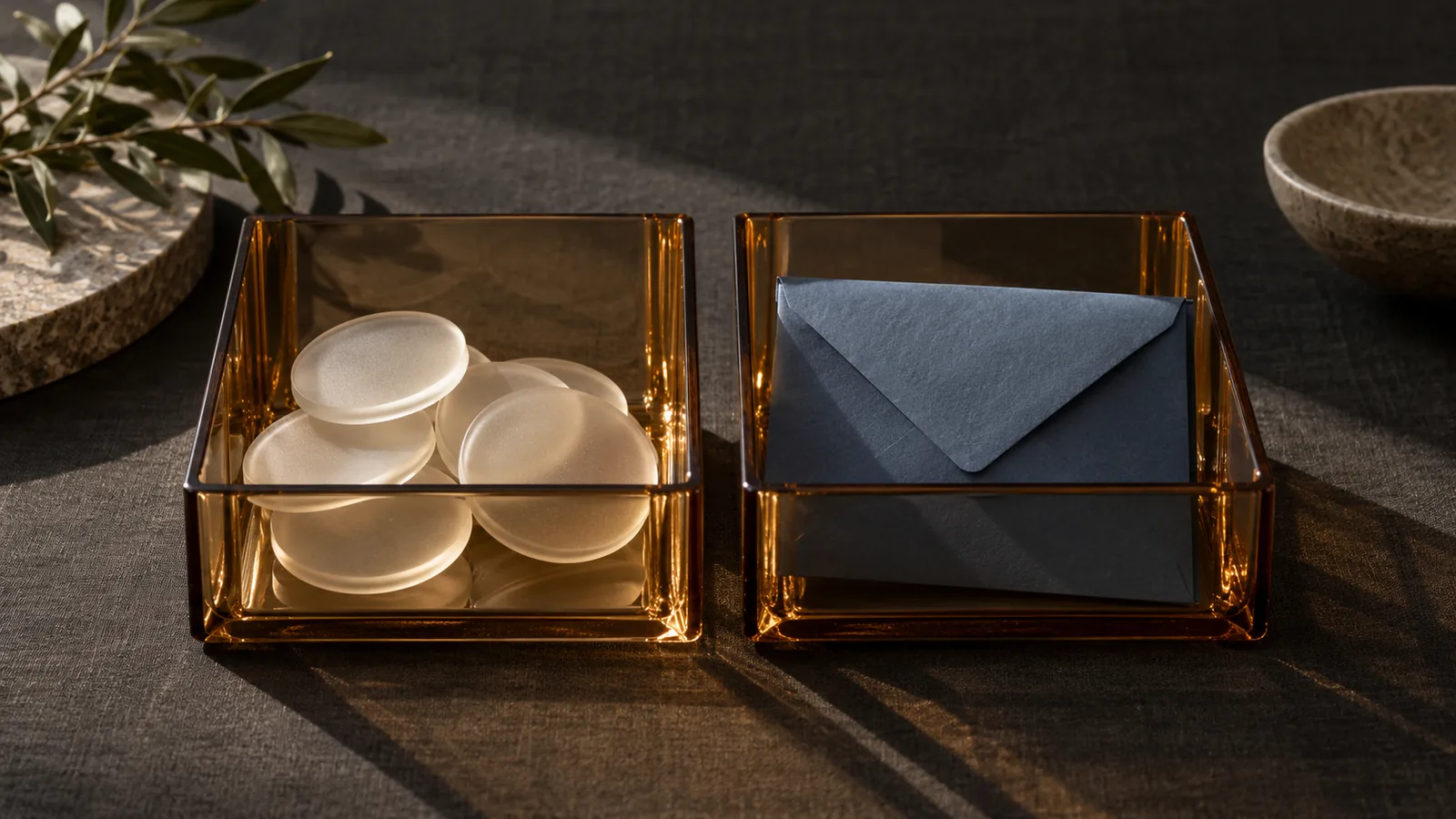Separate amber glass trays holding frosted discs and a folded blue envelope on dark linen